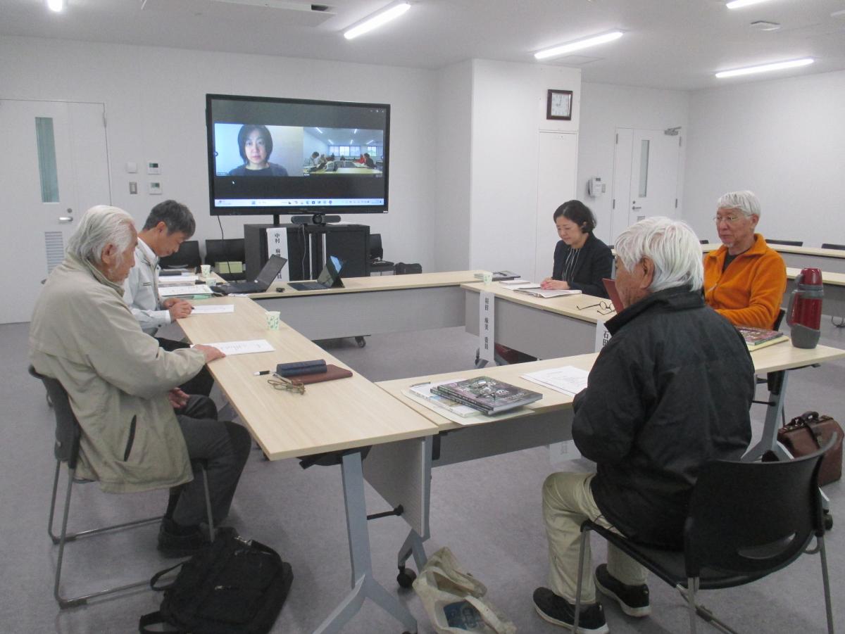 関係者達がロの字型に配置されたテーブルを囲んで座り、テーブル中央の大型モニターにはオンライン会議の参加者が映し出され会議が行われている自然部会の様子の写真