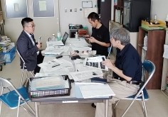 メガネをかけたスーツ姿の男性と2人の男性が向かい合って座り、大量の書類が広がる長机を囲んで話し込んでいる様子の写真