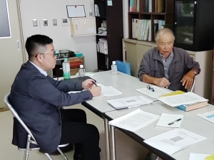 ダークスーツの男性と、グレーの上着を着た男性が、資料の置かれた白いテーブルを挟んで向かい合って座り、話している写真