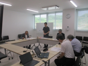 会議室で、黒い服を着た男性が奥のホワイトボードの前に立ち、ロの字の形に並べたテーブルに座る4人の出席者に向かって話をしている写真