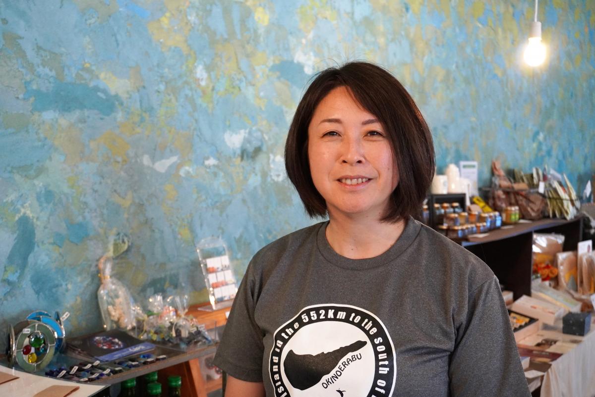 青と緑の色合いが混ざったテクスチャー壁を背景に、木製の棚に雑貨や食品が並ぶ店内でグレーのTシャツを着た女性が立っている写真