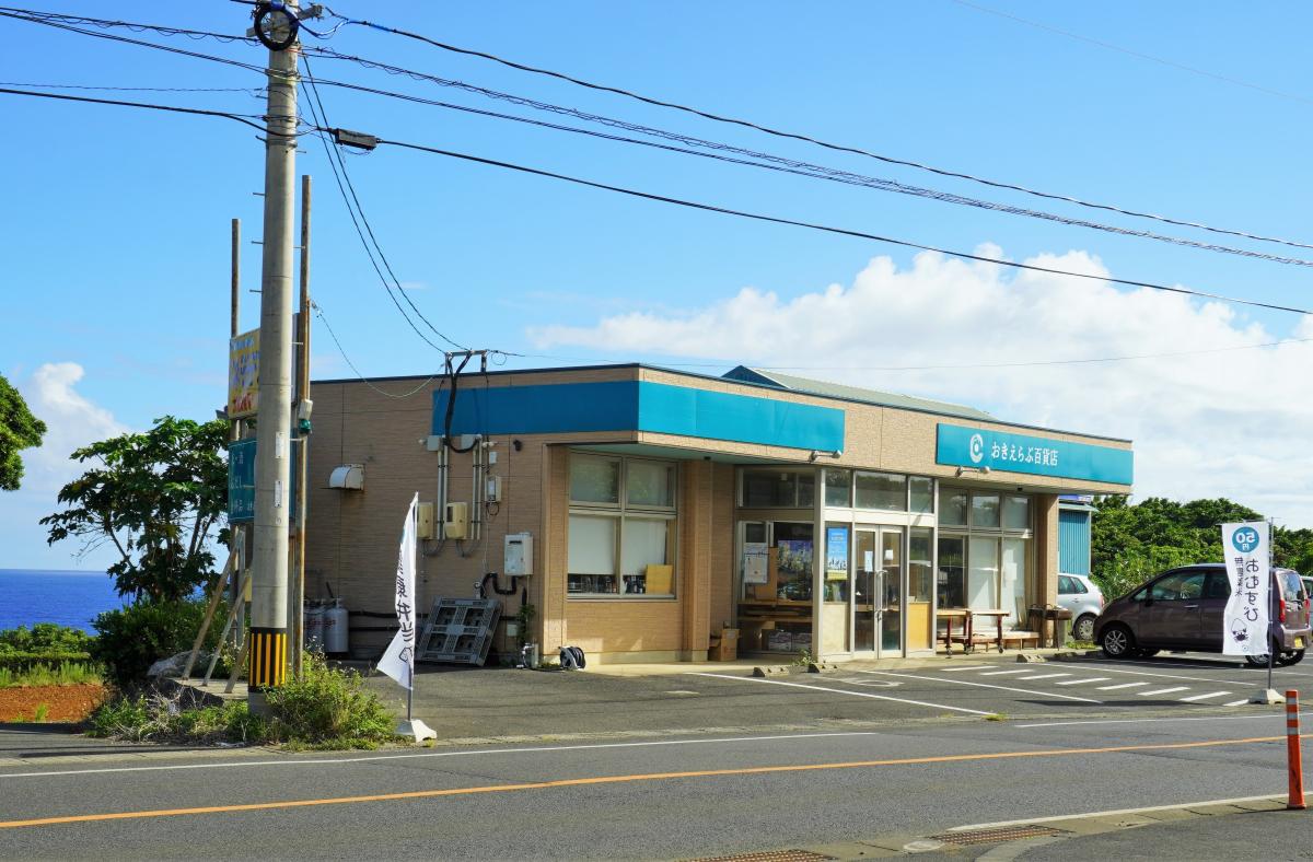 青空の下、海を背景に道路沿いに建つ平屋の店舗で、ベージュの外壁と青い屋根が特徴的な建物の前には駐車スペースがあり、白い旗に「おきのえらぶ」と書かれた文字が見える写真