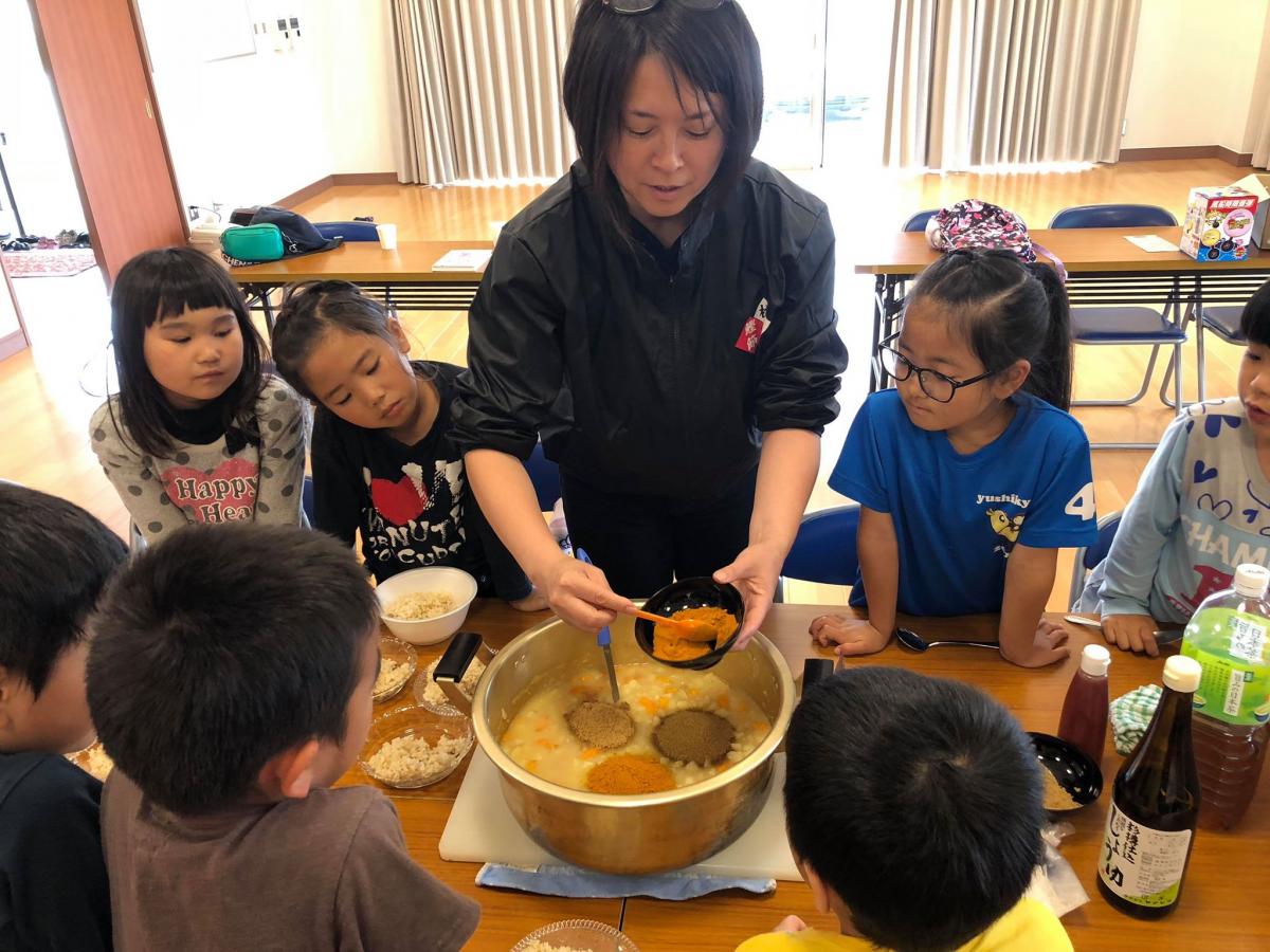 室内の長机に大きな鍋が置かれ周囲には子どもたちが座っており、中央にいる女性がカレーをよそっている料理体験の様子を写した写真