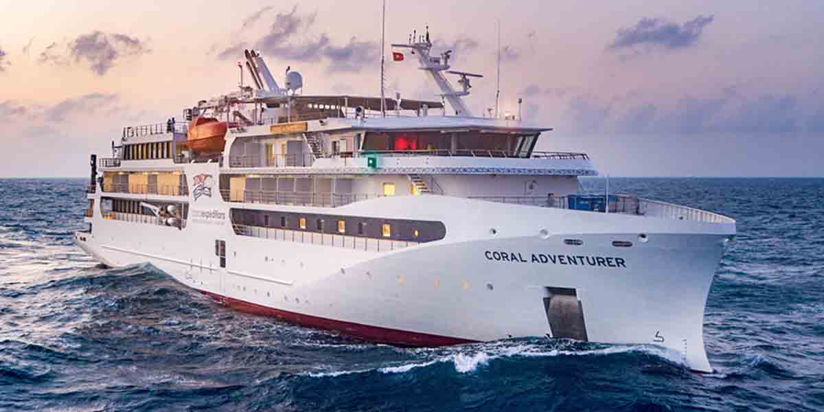 夕焼け空の下、波立つ暗い海原を進む白い船体に「CORAL ADVENTURER」と書かれた探検クルーズ船の迫力ある船首部分を捉えた写真