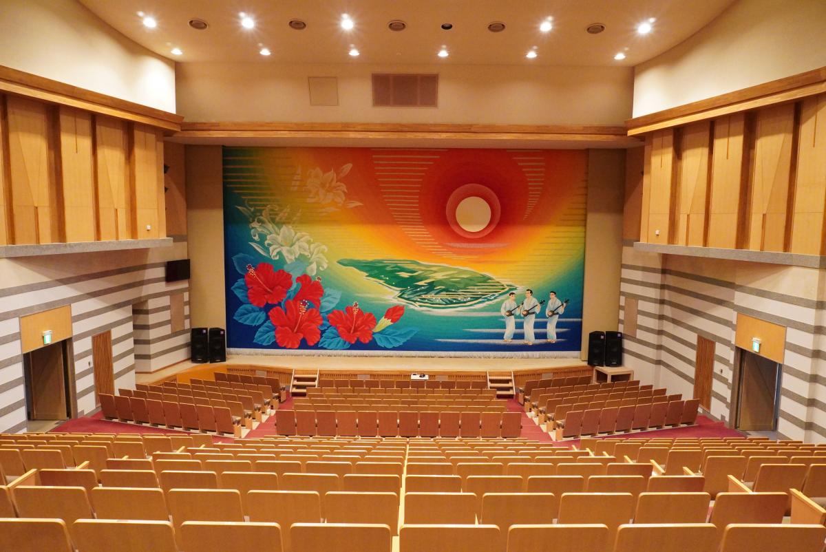 コンサートホールの舞台正面に壁一面に原色を多用したハイビスカスと白いユリなどの花々、太陽と海、そして三味線を持ち白い衣装を着た3人の人物が描かれたホールの緞帳と階段状に並んでいる客席がある文化ホール内を写した写真