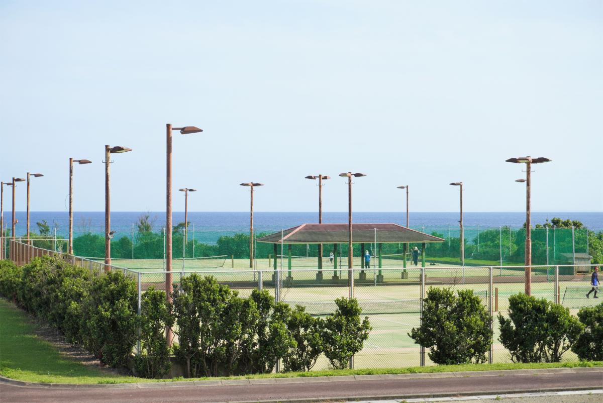 ナイター照明のポールが多数立ち並び、屋根付きの休憩所が中央にあるテニスコートの写真