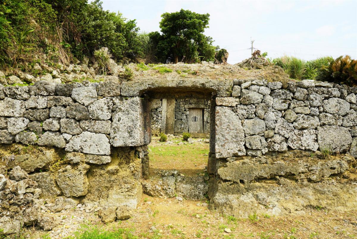 石積みの高い石垣の中央に四角い開口部の石門が設けられ、その奥に草地の中庭とさらに内側に石垣と木製の扉が見える遺跡の写真
