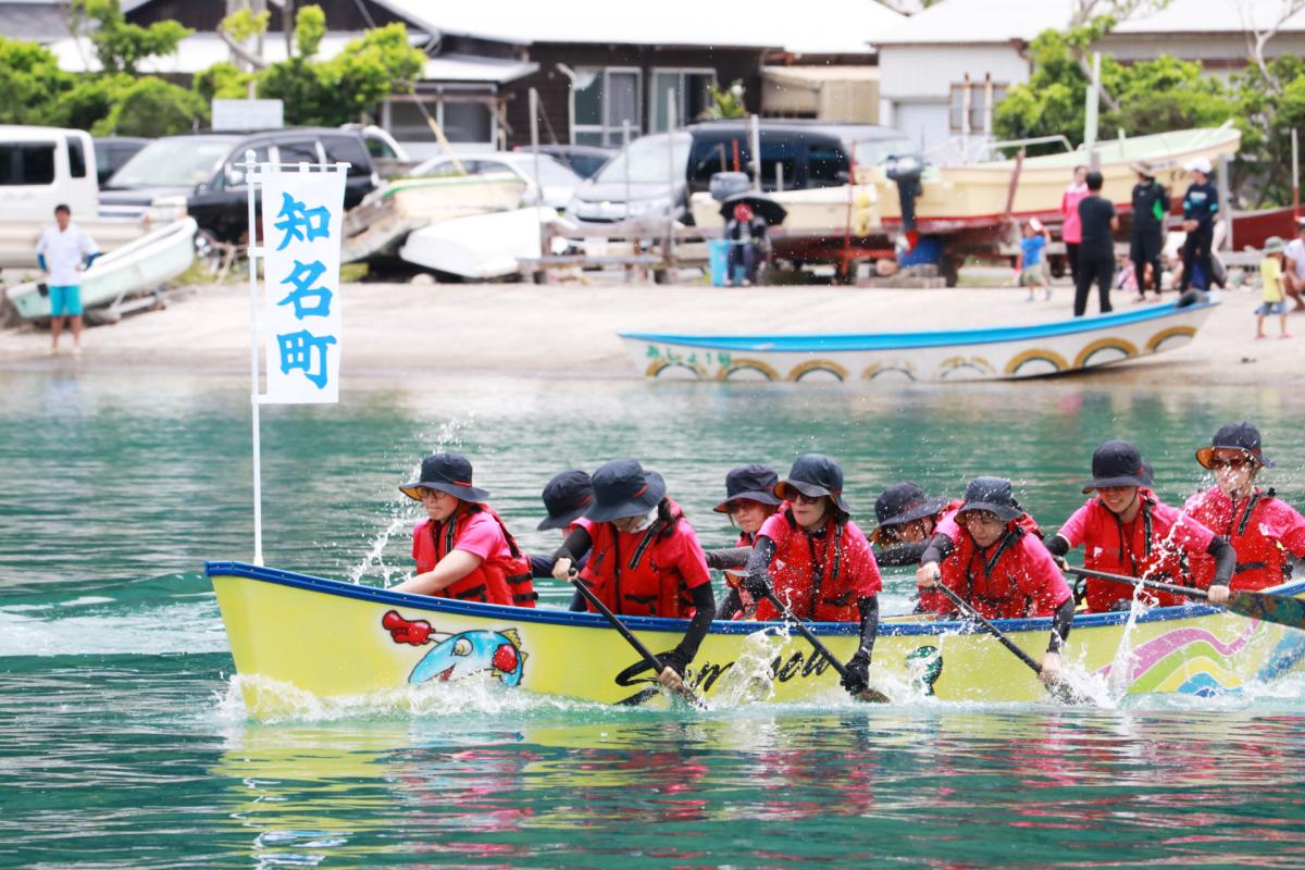 船首付近に「知名町」と書かれた旗が立っているくり舟に、赤いライフジャケットと帽子を着用した複数人の漕ぎ手が、息を合わせて力強くパドルを漕ぎ、水しぶきを上げながら進んでいるくり舟競争大会の様子の写真