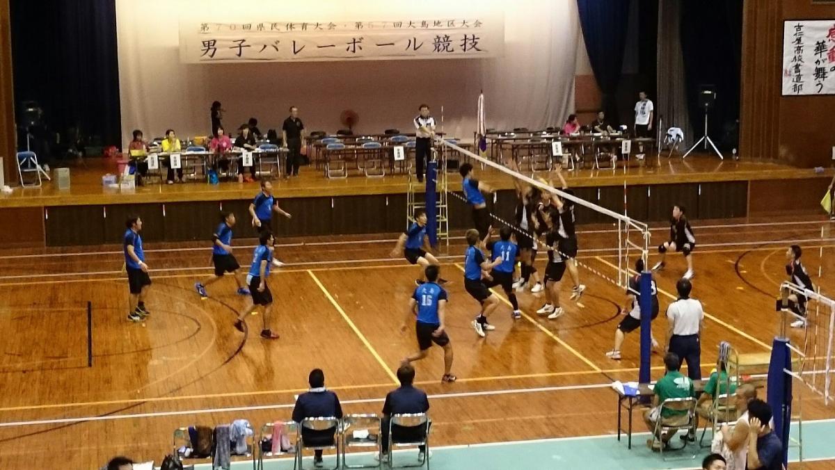 体育館で、青いユニフォームと黒いユニフォームに分かれた選手たちがネットを挟んで激しく男子バレーボールの試合をしており、コート外のテーブル席には審判や記録係と思われる人が座っているバレーボール競技の様子の写真