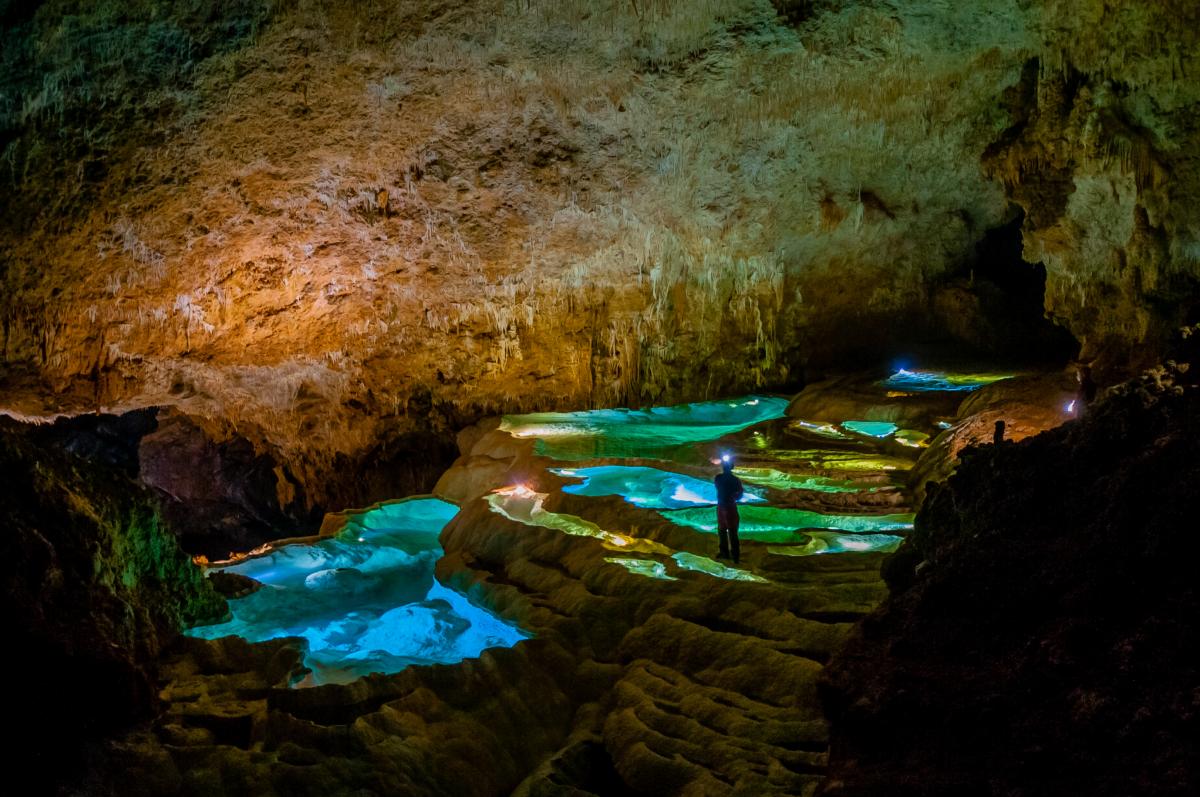 鍾乳洞の薄暗い洞窟内で、ヘッドライトを装着した人が立っている足元に、青く幻想的な光を放つ複数の斜面に形成される棚田のような水溜りが広がる写真