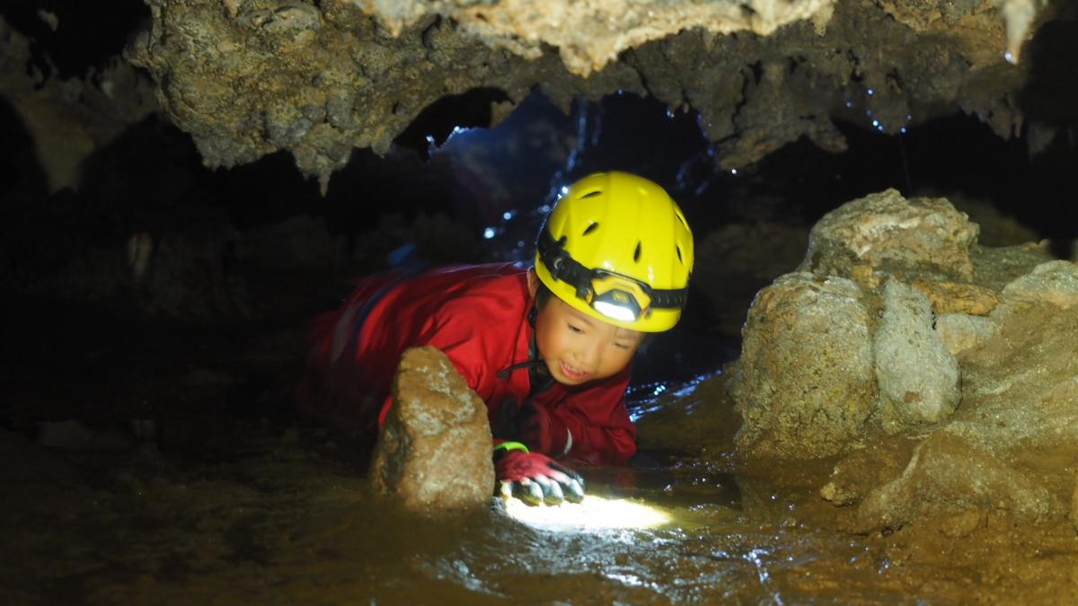 鍾乳洞の狭い通路を、赤いケイビングスーツと黄色いヘルメットを着用した子供が、水たまりの中を這って進む様子を捉えた写真