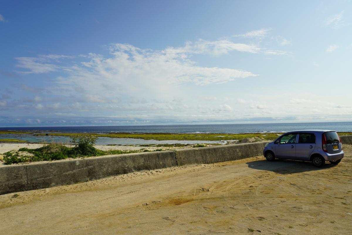 青い海と空、白い雲が広がる海岸線の手前にある砂利の広場に、レンタカーの軽自動車が駐車されている写真