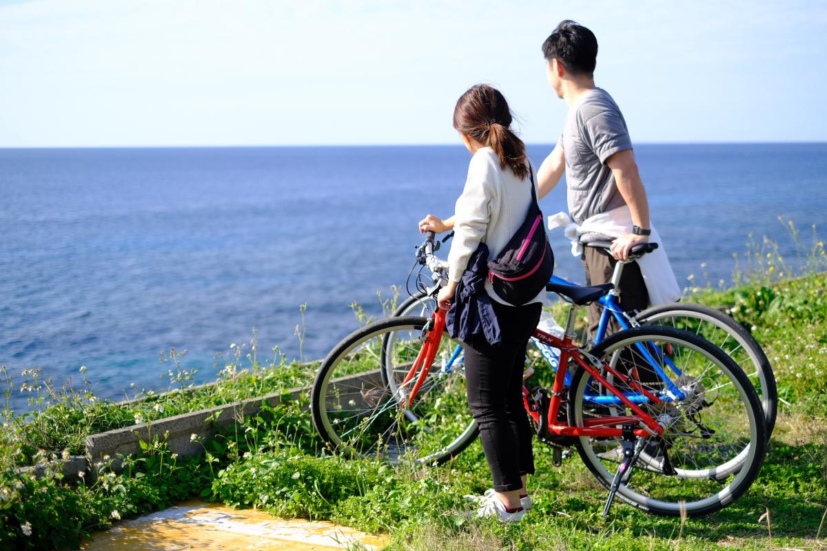 海が広がる草地の高台で、男女二人が赤と青の自転車を押しながら立ち止まり、水平線の見える青い海の方を並んで眺めている様子が写っている写真