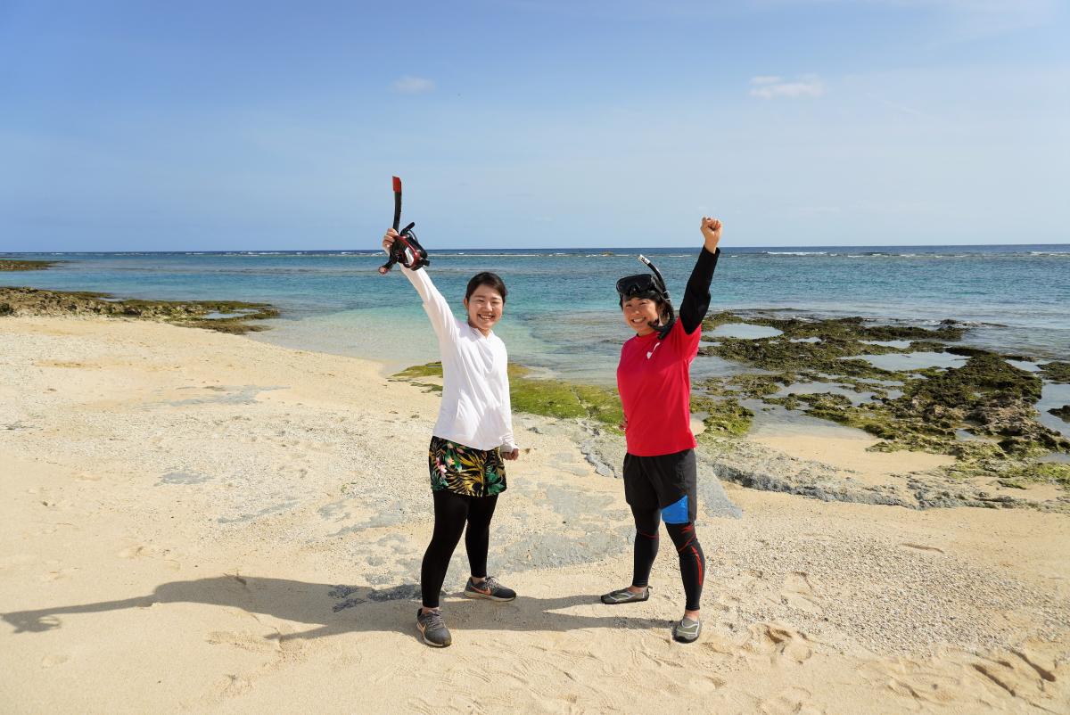 快晴のビーチで、シュノーケルを手にした女性とシュノーケルを付けた女性が海を背景に笑顔で右手を上げている写真