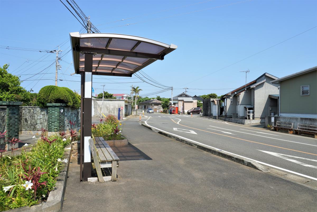 屋根付きの木製ベンチが置かれたバス停から、緩やかにカーブする道路の先に民家や商店が並ぶ集落の風景を見渡した、のどかで開放感のある田皆小学校区の風景写真