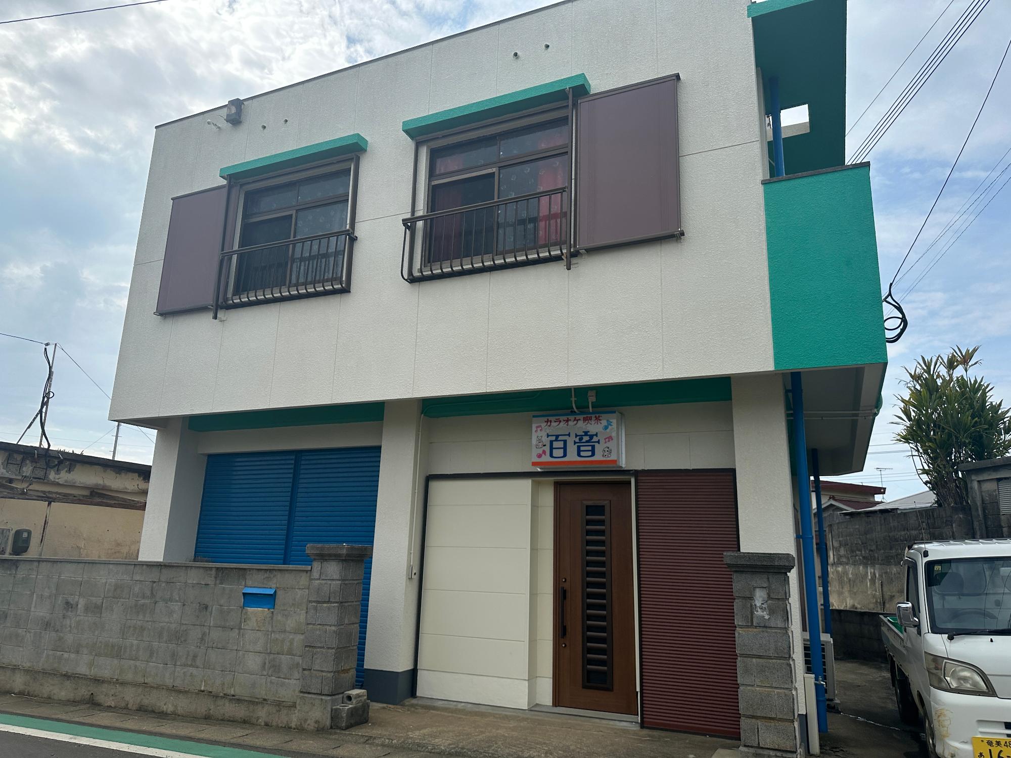 物件写真 白い壁の2階建ての建物が、青と茶色の雨戸や縁でアクセントがつけられ、玄関上には「カラオケ喫茶 百音」と書かれた看板があり、その前には低いブロック塀がある物件No.097の外観写真