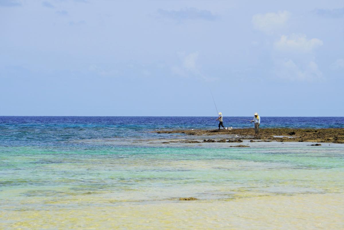 澄んだターコイズブルーの浅瀬が広がり、沖合の濃い青色の海との境目に露出した茶色い岩礁に、二人の男性が立って釣りをしている様子の写真