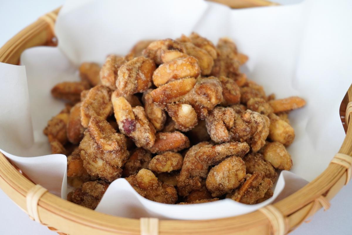 砂糖や飴でコーティングされた茶色く光沢のあるピーナッツやナッツのような豆菓子が、白い紙を敷いた素朴な編み込みの丸い竹かごに山盛りにされた写真