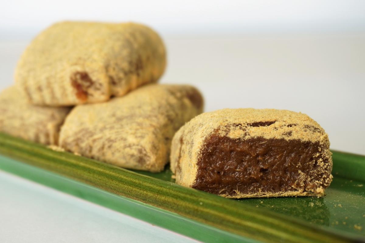 きな粉がまぶされた四角い田芋もちが縦長の艶やかな緑の皿に盛られ、右奥は田芋もちが3つ重ねられ手前のものは中身が見えるように切られて添えられた写真