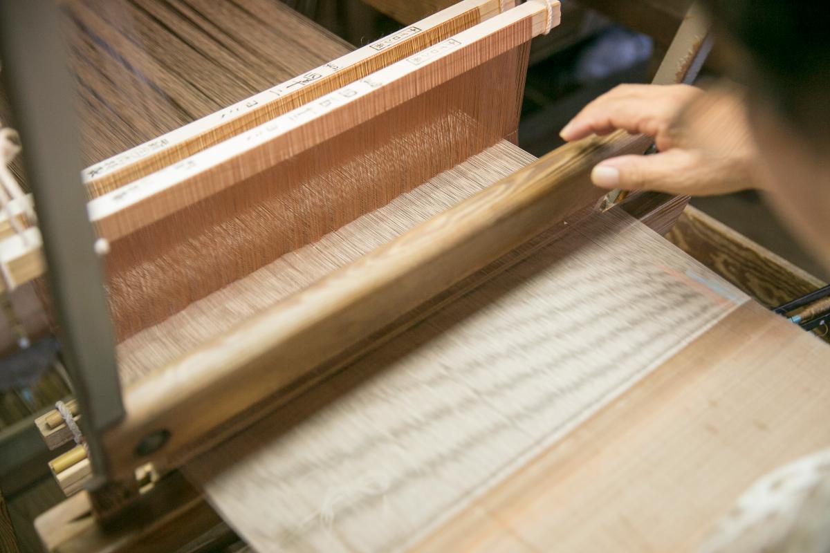 織機の一部と、縦糸が張られた状態で茶色がかった繊細な糸を織るために、手前で木の棒を操作している職人の手を、肩越しに捉えた写真