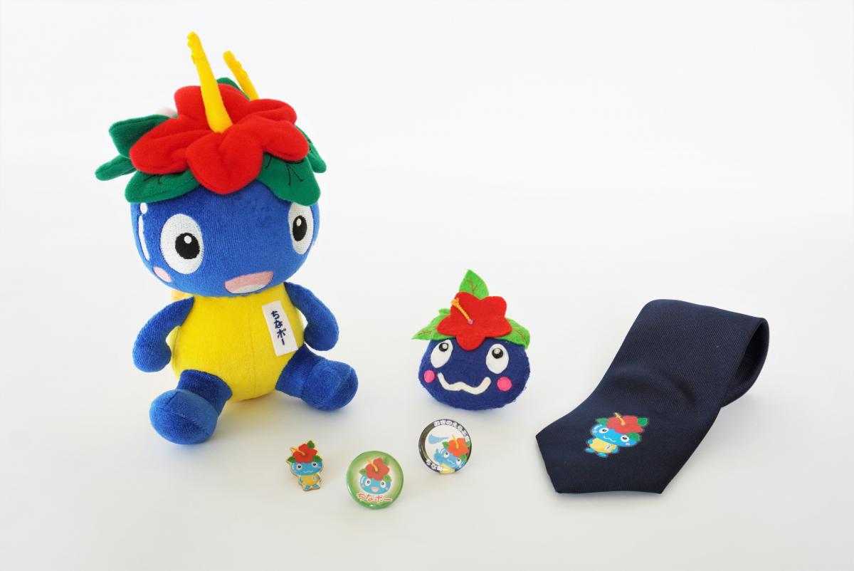 青い体に黄色の腹部、頭にハイビスカス赤い花と緑の葉、黄色い角を持つちなボーのぬいぐるみと、小さなマスコット、バッジ、ちなボーの刺繍が入った紺色のネクタイが置かれた写真