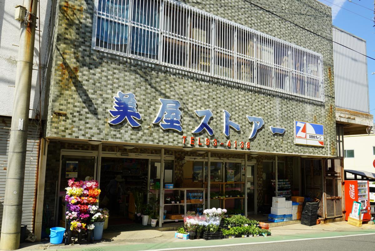 黄土色のタイル張りの外壁を持つ二階建ての「美屋ストア」の正面が捉えられ、軒先には生花や鉢植え、商品などが並んだ写真