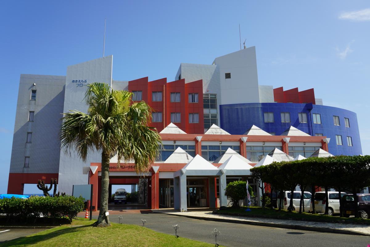 手前右に車が並ぶ駐車場と剪定された生け垣があり、白い三角屋根が並ぶ正面入口の背後に、青い円弧状の外壁が組み合わさった、赤・白の色鮮やかな外観が特徴的な「おきえらぶフローラルホテル」の外観写真