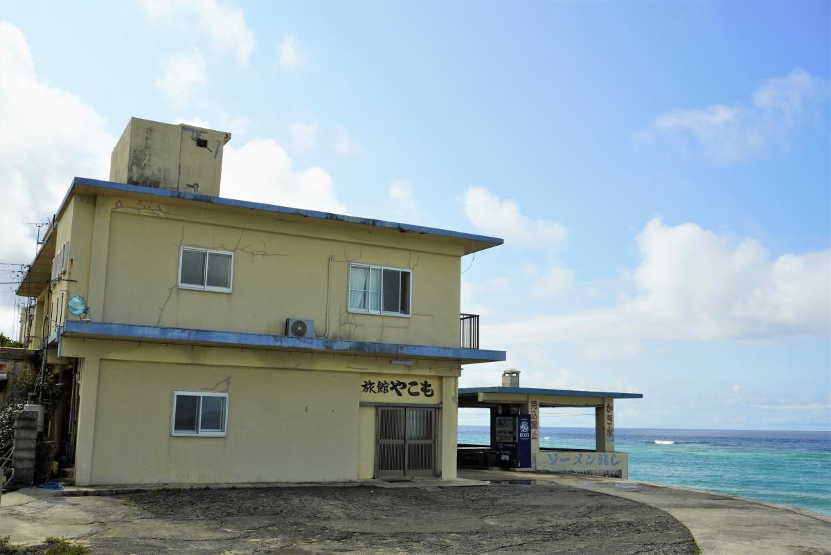 海沿いにあり、黄色い外壁と青い縁取りが特徴的な二階建ての建物で、後方にきれいな海が見えている旅館やこもの外観写真