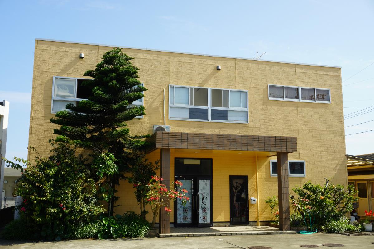 黄色い外壁と焦げ茶色の庇が特徴的な二階建ての建物で、手前には緑の植え込みがある民宿松屋の外観写真