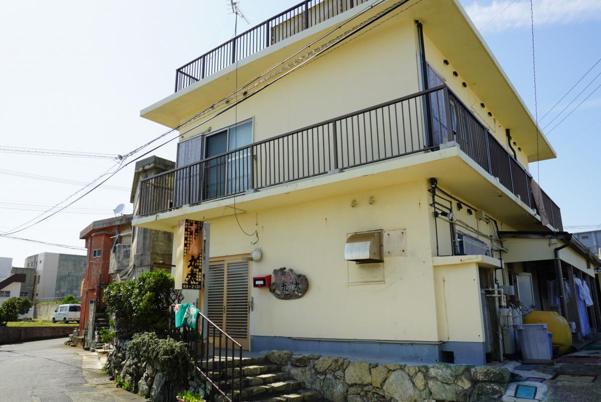 二階に金属製の手すり付きバルコニーを持つ淡いクリーム色の外壁の民宿寿司・丸太の外観写真