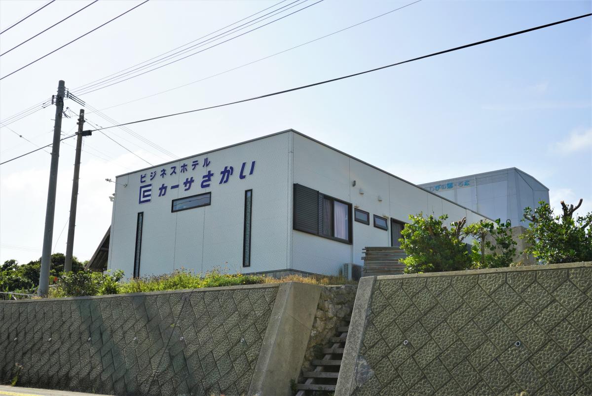 白いプレハブ構造で、上部に「ビジネスホテル E カーサさかい」と青い文字で書かれたシンプルなホテルを、手前の石積みと階段越しに斜め前方から捉えたビジネスホテル カーサさかいの外観写真