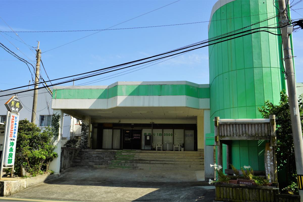 白い外壁にライトグリーンに塗装された正面玄関があり、建物の右側には円筒形のライムグリーンの塔状の部分があり、左側には看板が立っているパラダイスINN沖永良部の外観写真