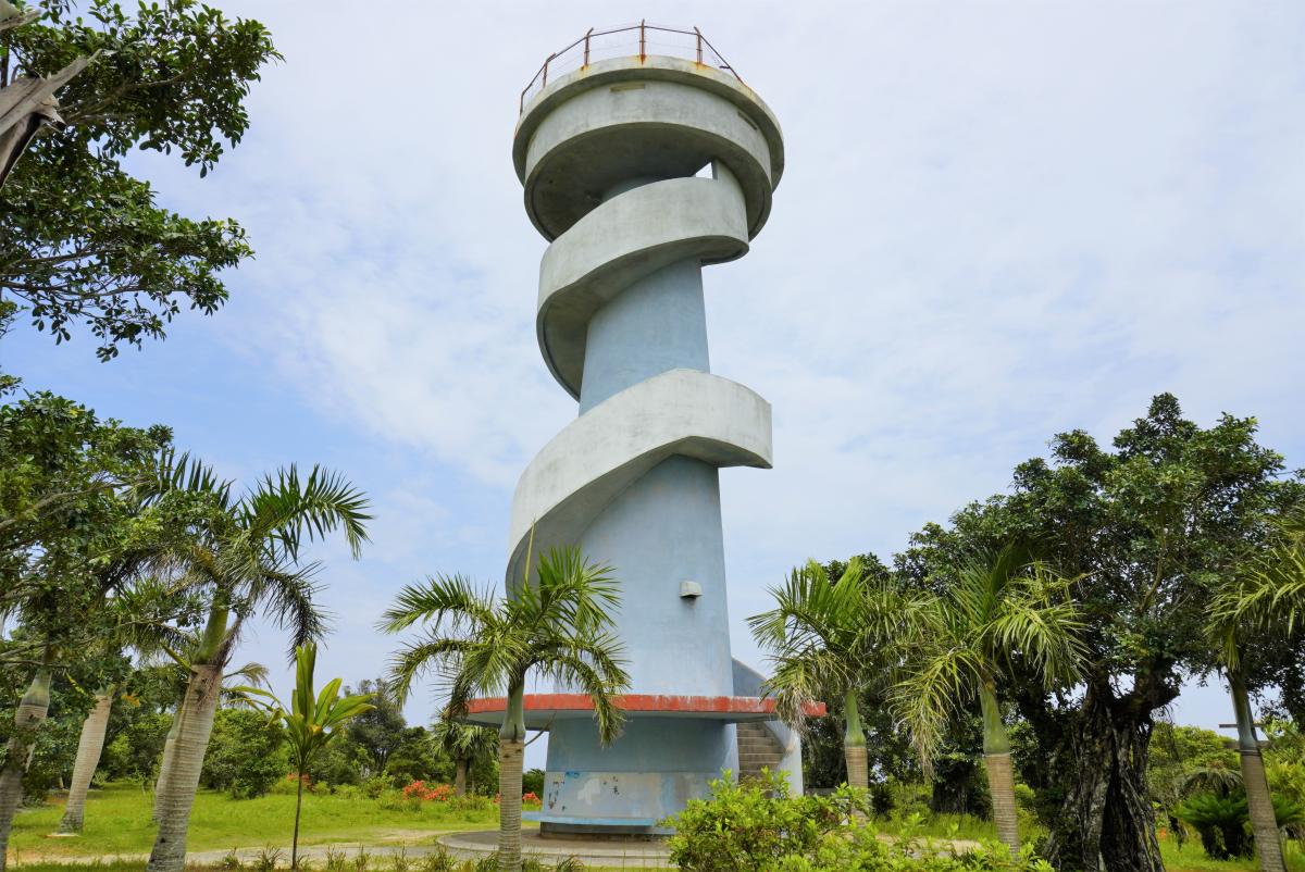 円筒状の展望台の外壁に沿って、青灰色のらせん階段が上部へ巻き付いており、下部はヤシの木を含む豊かな熱帯植物に囲まれている大山展望台の写真