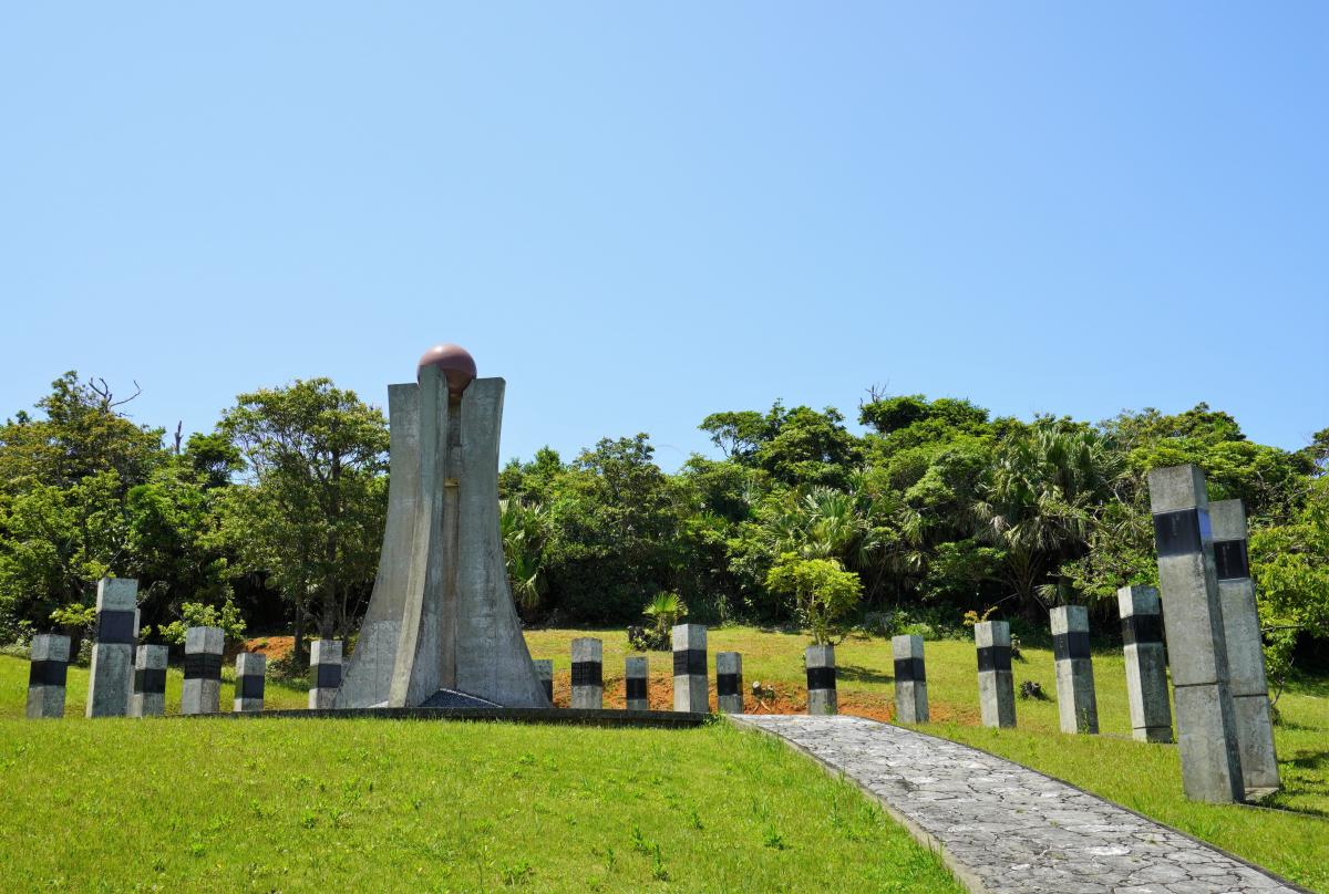 芝生の広場の中央にそびえ立つ、上部に茶色の球体が挟まれたコンクリート製のモニュメントと、その周りを囲むように直立した石柱が配置され、奥には豊かな緑の樹木が茂り、モニュメントへと続く石畳の歩道が手前から奥へ伸びる知名町平和之塔の写真
