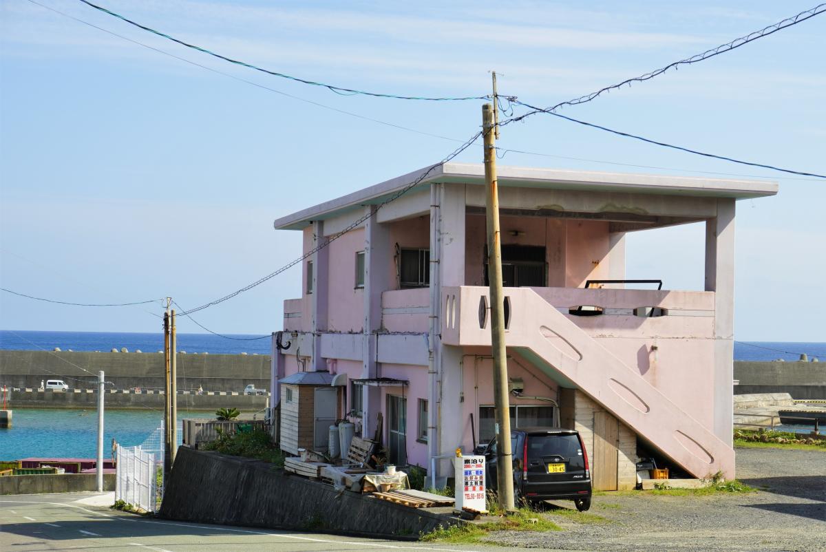 奥には青い海と灰色の防波堤が見える場所に立つ、ピンク色の壁の二階建ての建物のビジネスワンルーム潮風の外観写真