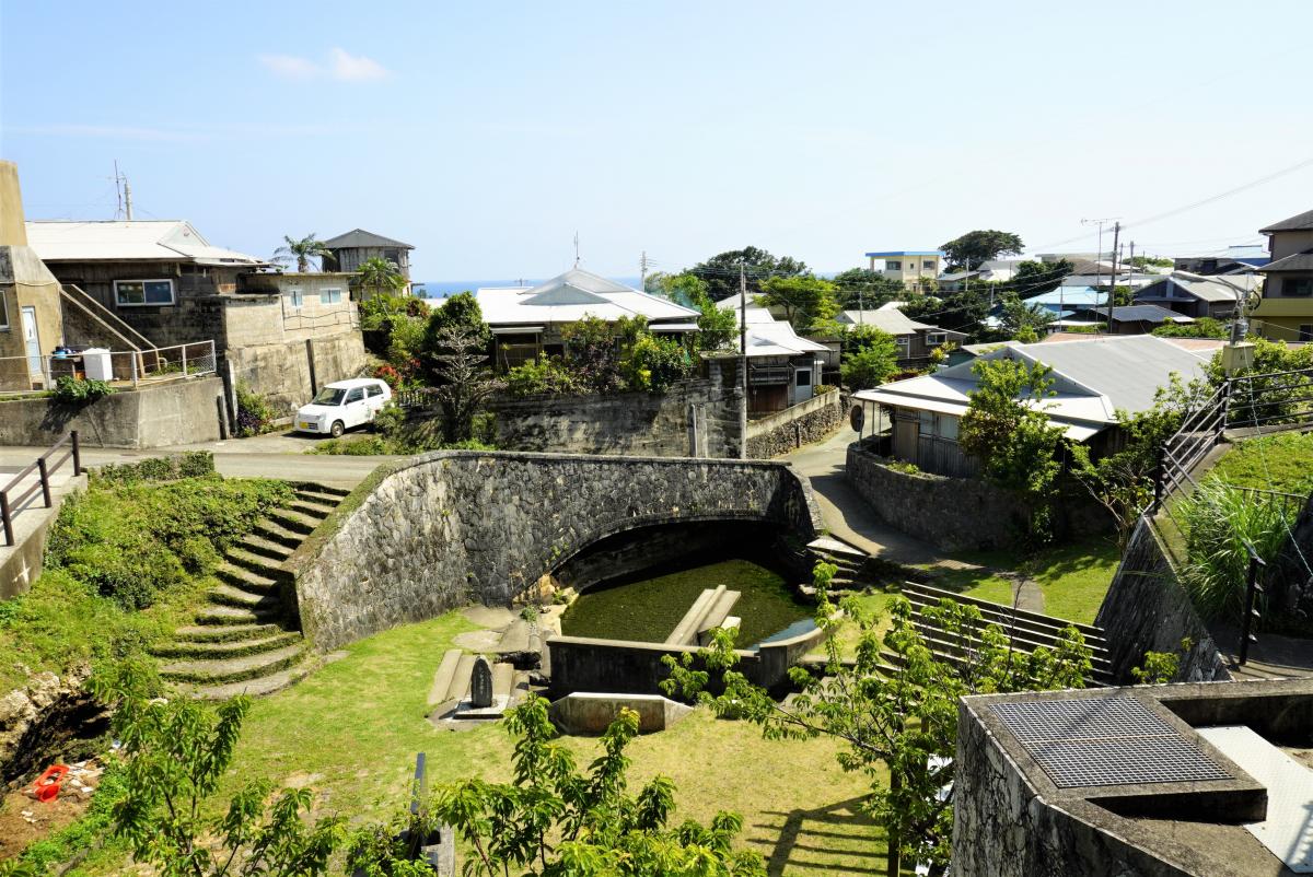 青空の下、石造りのアーチ状の橋と、その下に水たまり、周囲には石の階段とコンクリートの構造物、その奥には民家の建物が建ち並ぶ集落の風景を捉えた写真