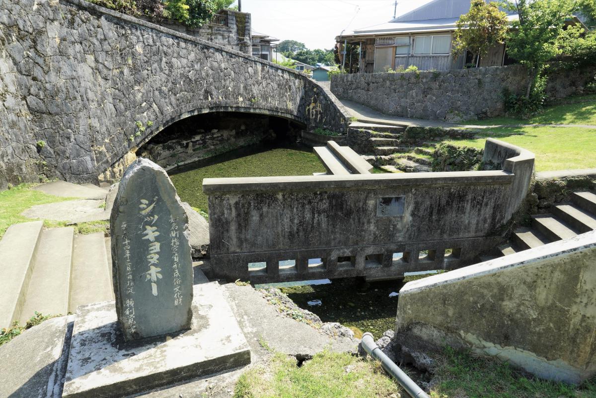 石積みのアーチ状の橋が架かり、その手前を古いコンクリートの短い橋がまたぎ、手前左下には「ジッキョヌホー」と刻まれた石碑が立つ、石垣に囲まれた水辺の風景写真