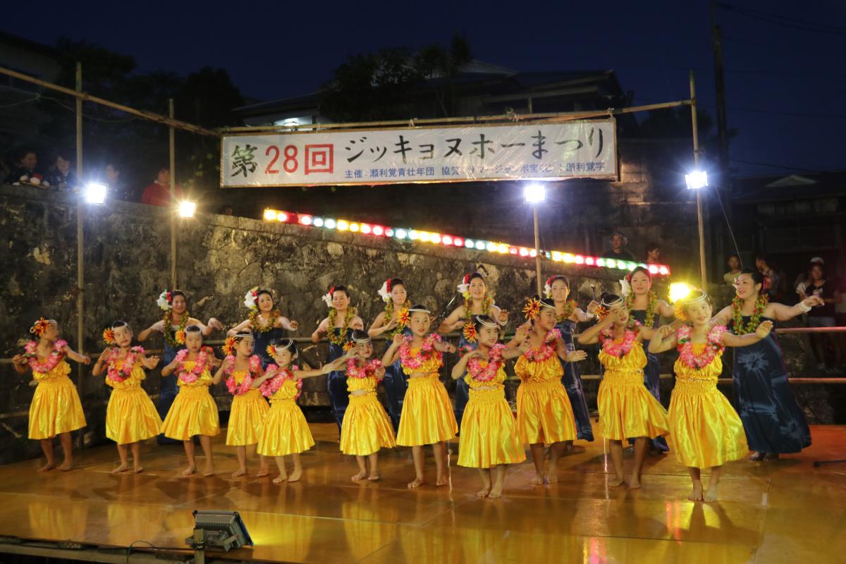 夜の屋外ステージで、黄色い衣装にハイビスカスのレイを身につけた少女たちと、紺色の衣装の大人たちが、華やかな照明の下でフラダンスを踊っている写真