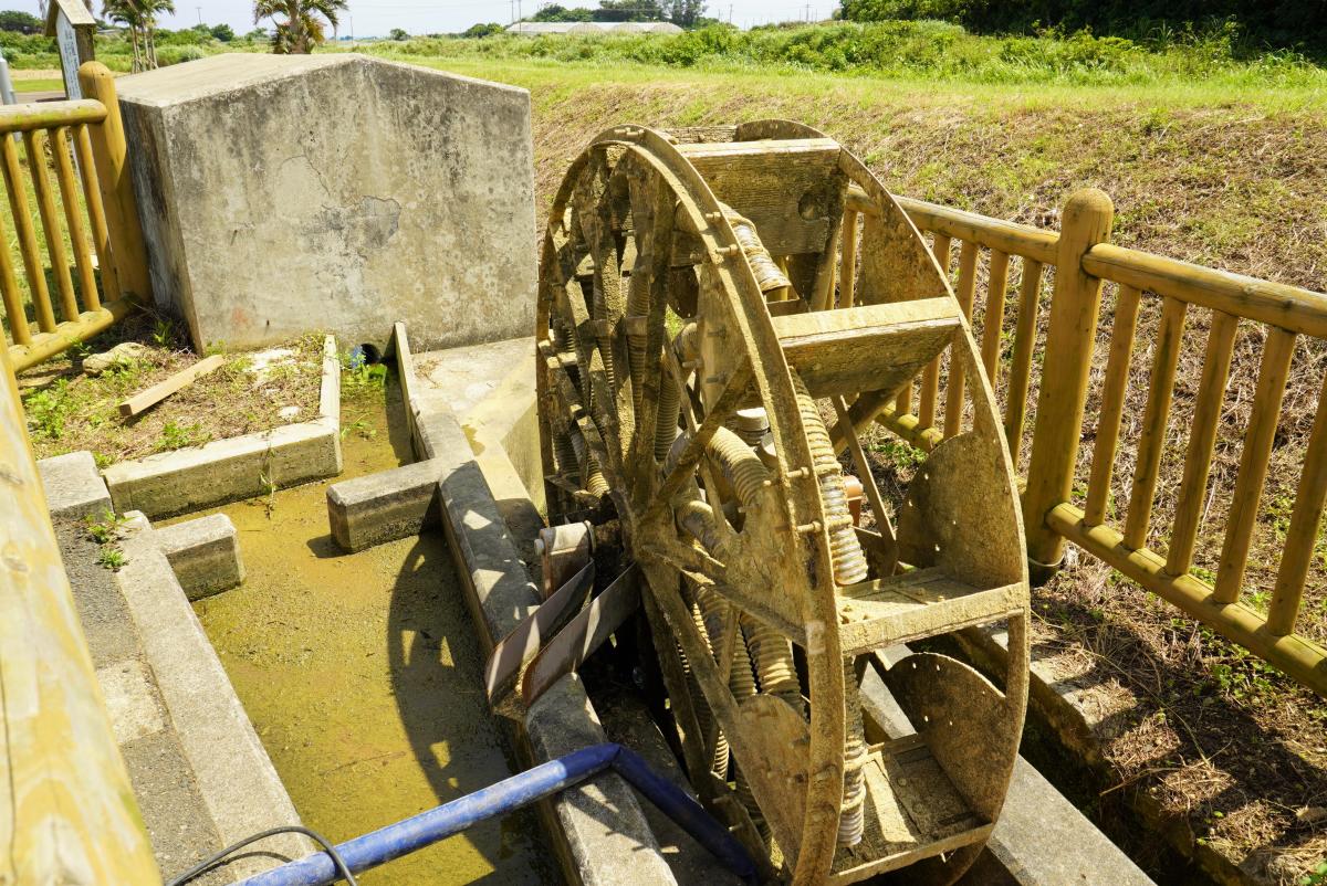 水路の脇に木製の水車とコンクリートの構造物があり、水車は一部が水に浸かり、周囲は木製の柵で囲まれている写真
