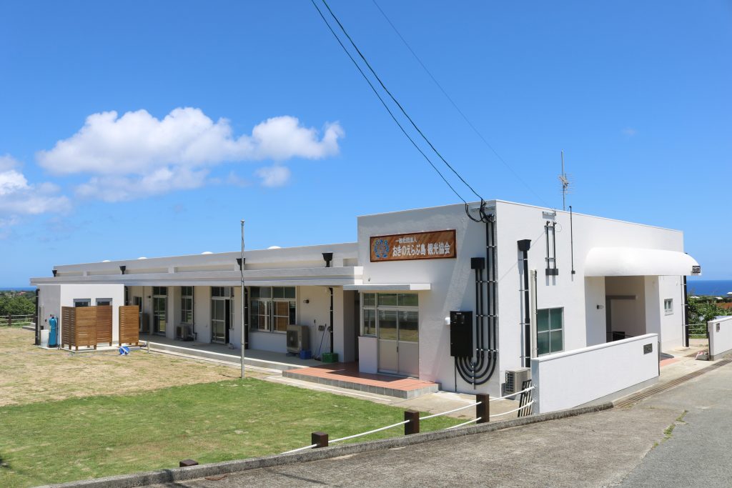 白い平屋建ての建物が青空と海を背景に立ち、正面にはエラブココの目印である「地域のふれあい福祉施設」と書かれた看板があり、広い芝生の庭と舗装された通路が整備された写真
