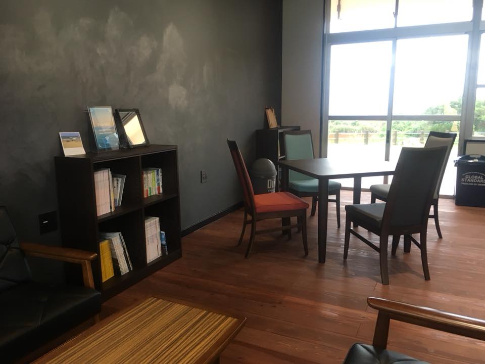 黒い壁と大きな窓に囲まれたカフェスペースには、木製の床にソファとテーブルが置かれ、奥には本や写真立てを並べた本棚と複数の椅子がある落ち着いた雰囲気の写真