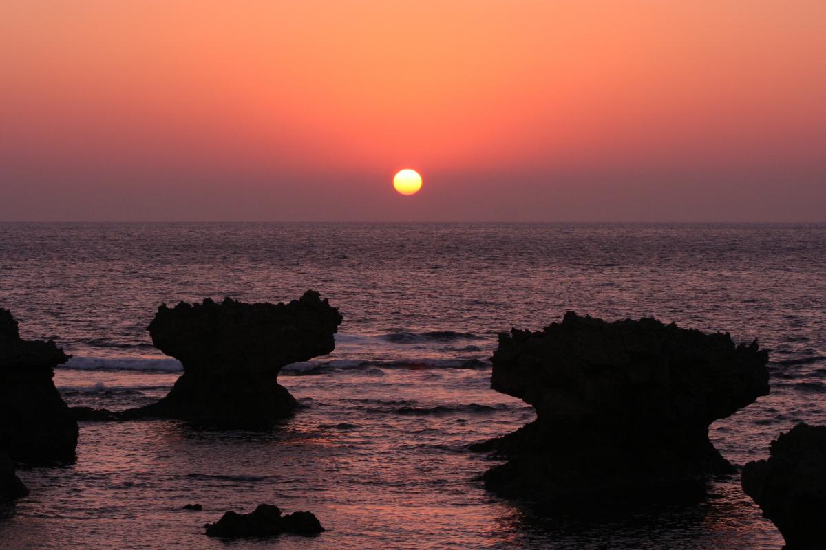 空と海が濃いオレンジ色に染まる中、海面から奇岩のシルエットが浮かび上がり、水平線上に真ん丸でまばゆい太陽が輝く情景写真