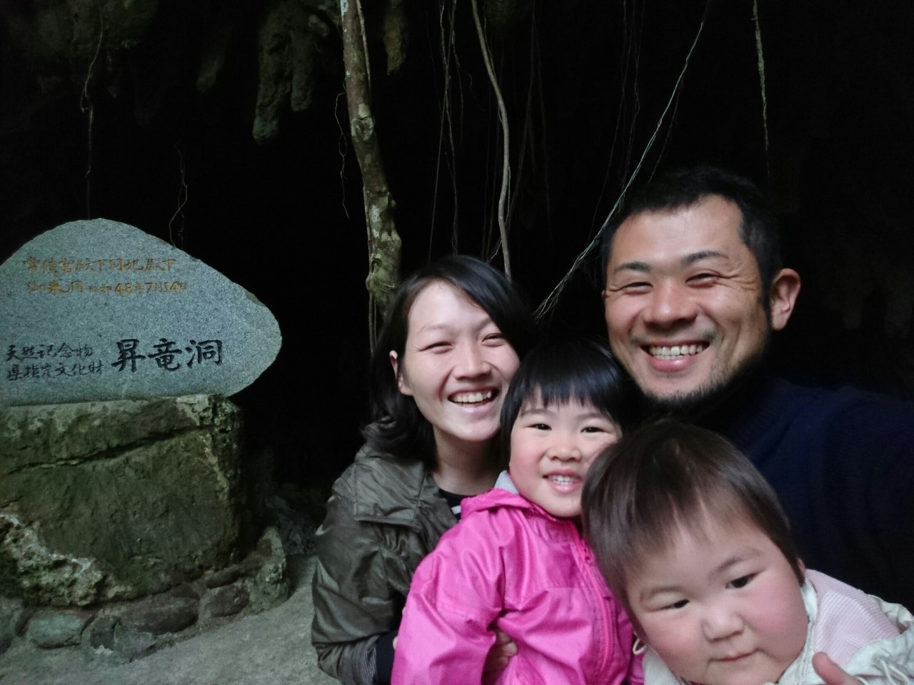 昇竜洞と刻まれた石碑が置かれた暗い洞窟の入口前で、勘里さん一家4人が笑顔で記念撮影をしている写真