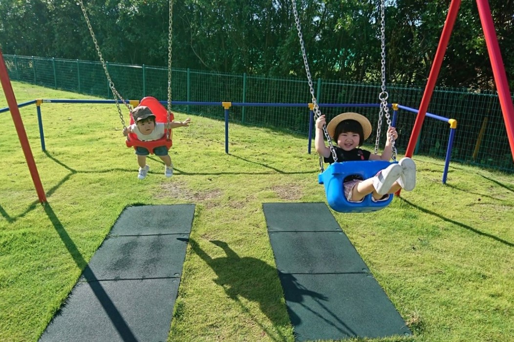 緑の芝生に囲まれた明るい屋外の遊具エリアで、赤と青の座席を備えた二つのブランコに帽子を被った子どもが元気に遊んでいる写真