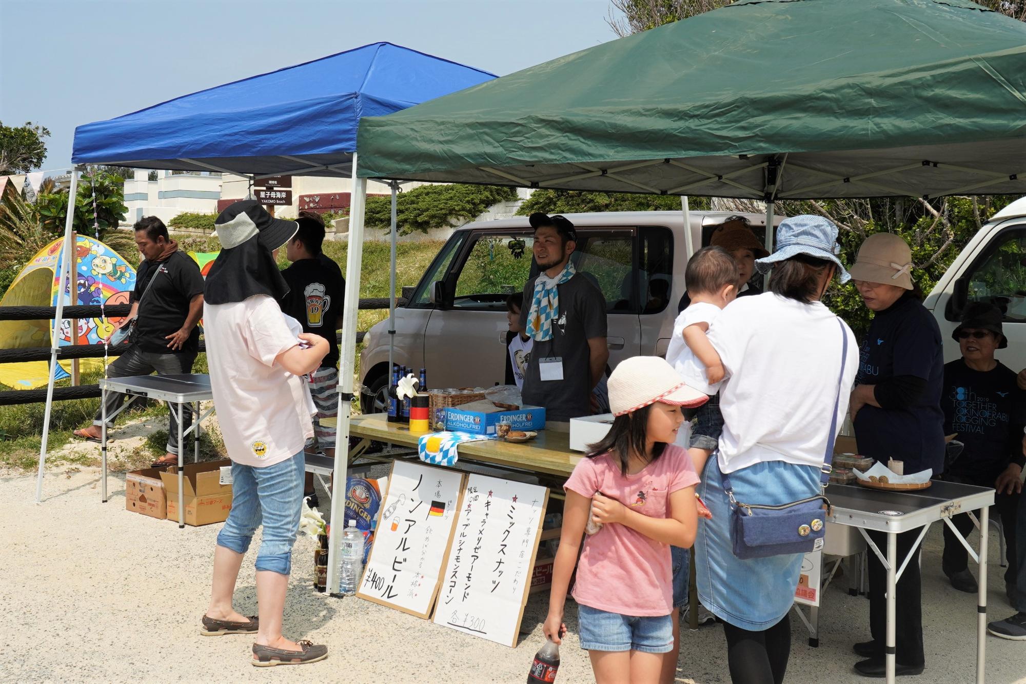 屋外のイベント会場で、青と緑のテントの下にテーブルが並び、ノンアルコールビールやミックスナッツなどのおつまみが書かれた看板が立てられ、店員とお客さんが談笑している写真