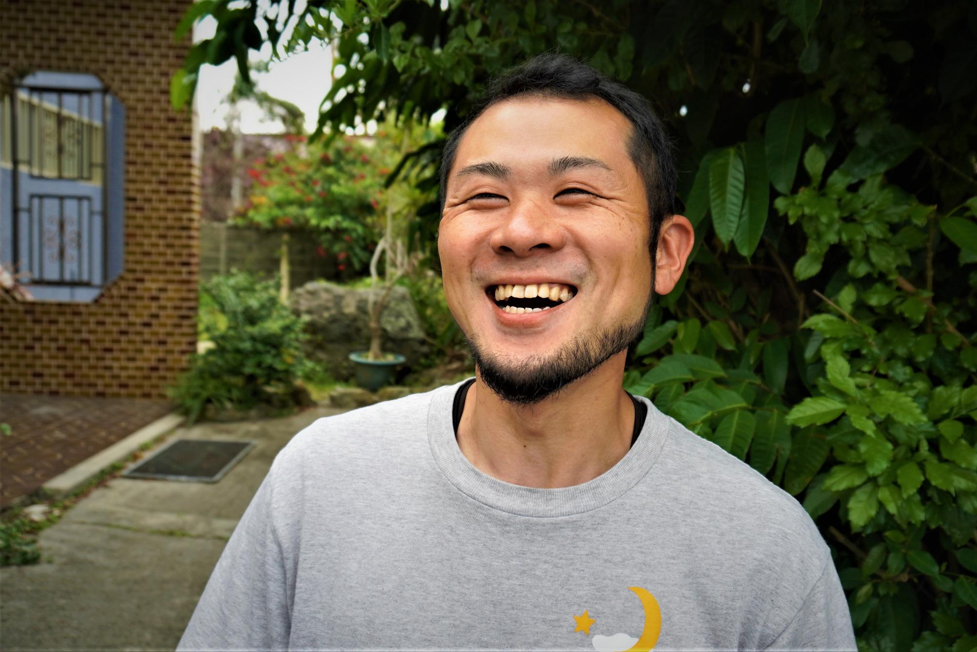 灰色のTシャツを着た勘里さんが屋外に立っており、背後にはレンガ造りの建物と舗装された地面、緑豊かな庭木が見える写真