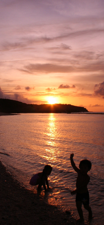 オレンジ色に染まる空の下、太陽が海面に光の筋を作り出しており、波打ち際では二人の子供のシルエットが楽しそうに遊んでい様子を写した海岸風景の写真（空編のページへリンク）