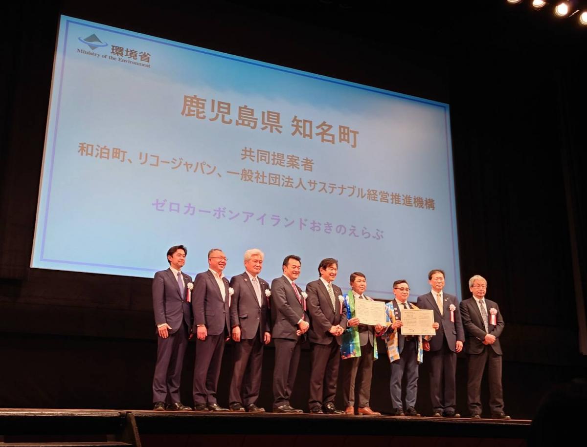 環境省のロゴが表示された大きなスクリーンを背景に、鹿児島県知名町のゼロカーボンアイランド推進に関する表彰式典で、複数のスーツ姿の人物が並び、中央の人物が脱炭素先行地域選定証を掲げている写真。