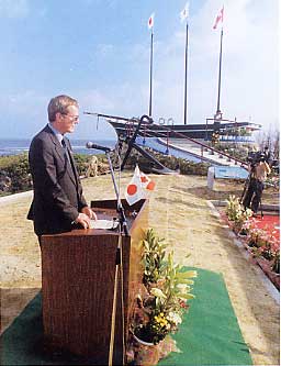 青空の下、レナード・J・エドワーズ駐日カナダ大使が日本国旗の付いた演台でスピーチをしており、背景には3本の旗を掲げた黒い船のモニュメントが見えるトゥループ号110年祭の様子を写した写真