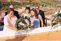 屋外で複数の乗組員の遺族が白い台の前に並び、緑の葉と白い花で飾られた花輪を献花している様子が写されている写真